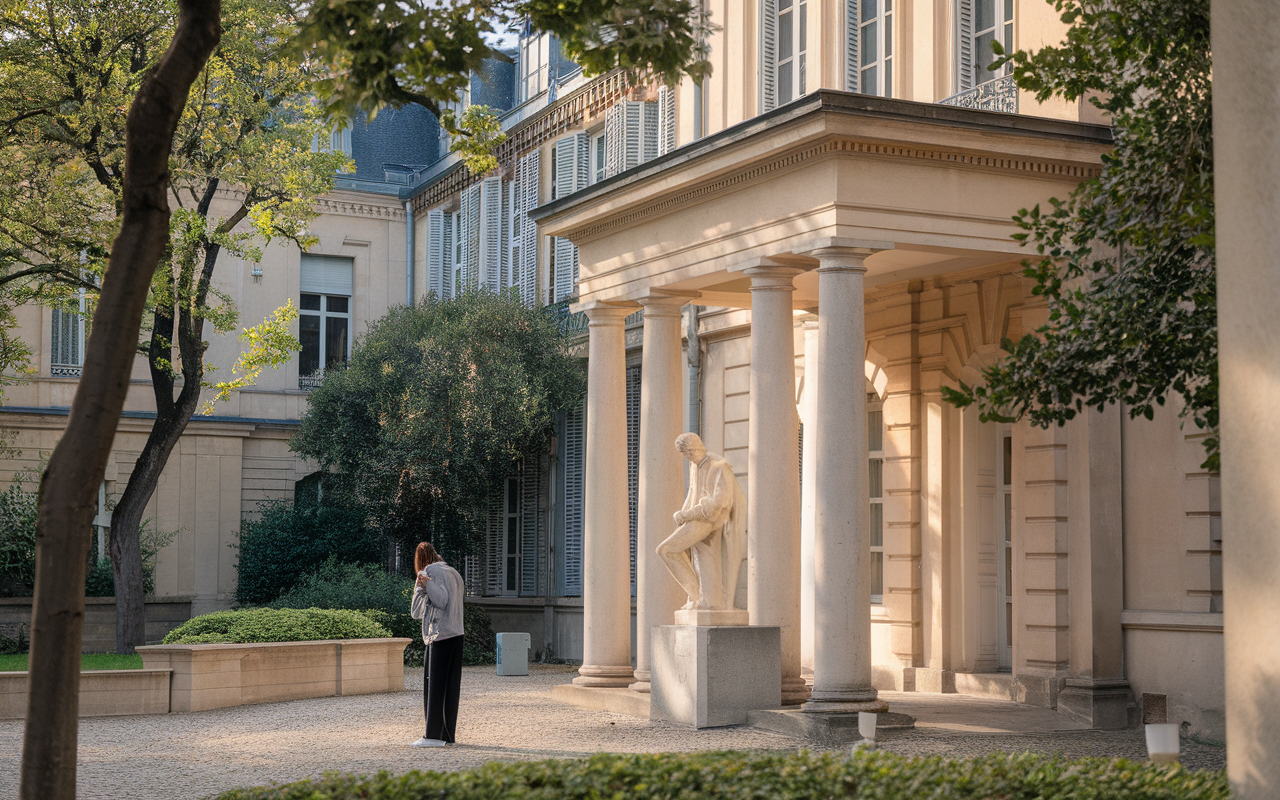 Une femme se tient près d'une statue en marbre, devant un bâtiment élégant avec des colonnes, entourée d'un espace verdoyant.