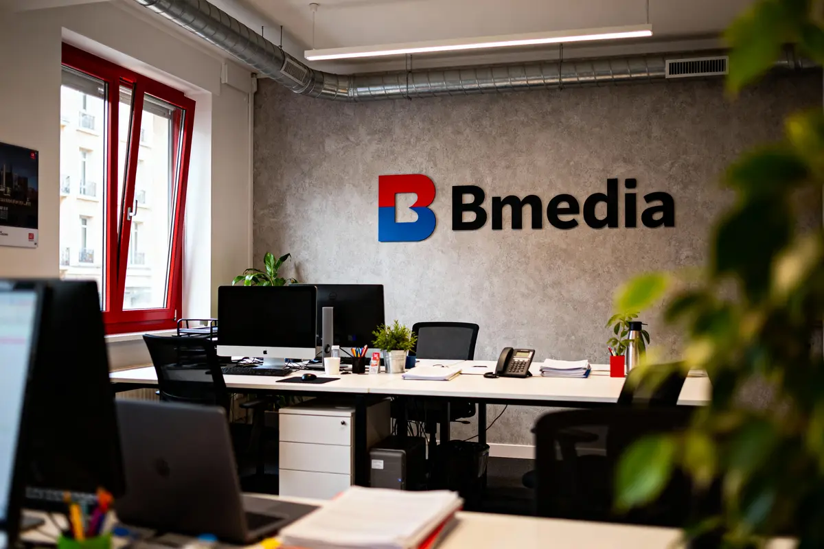 Bureau moderne avec murs gris, logo Bmedia, ordinateurs, plantes, et fenĂȘtres rouges.