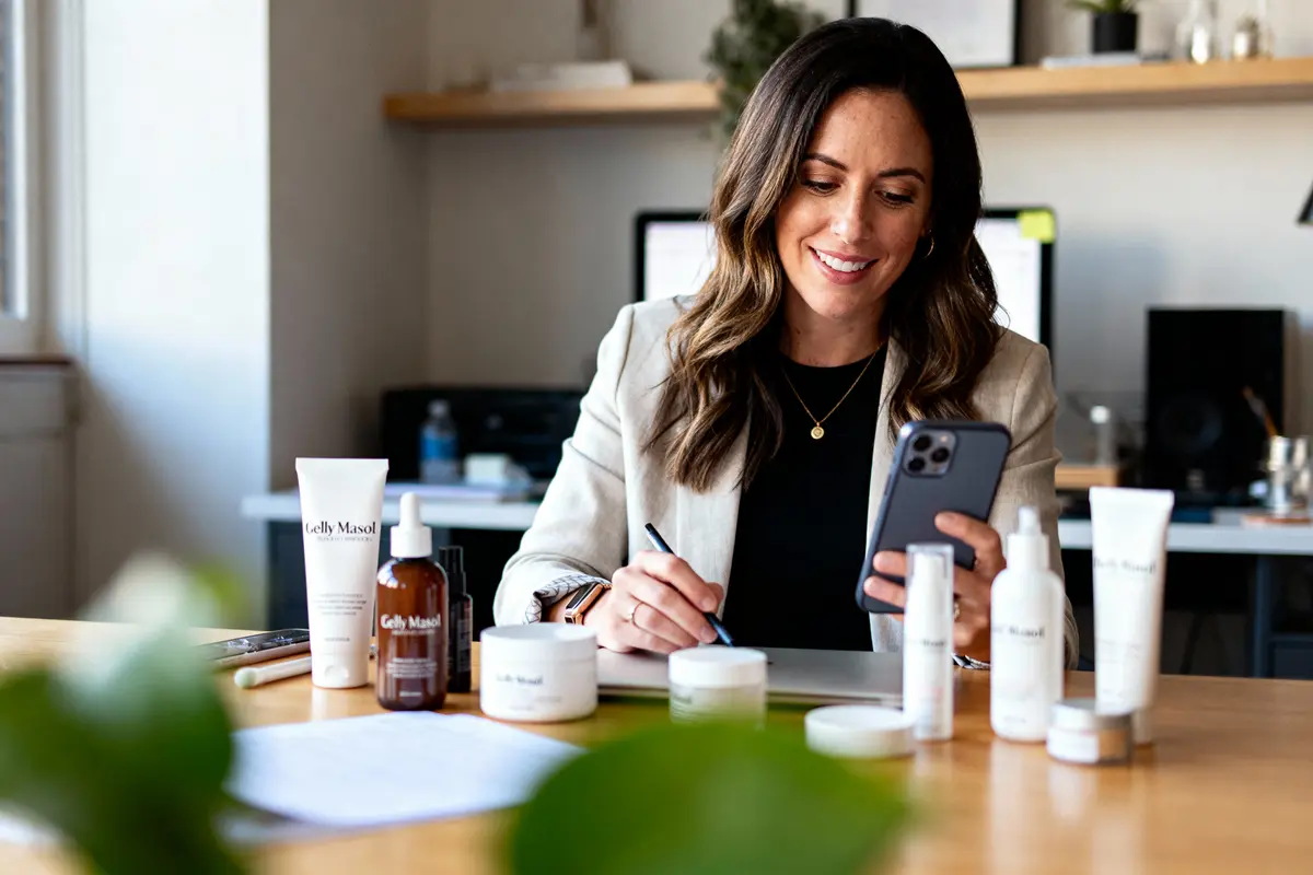 Femme souriante travaille au bureau avec téléphone, documents, et produits de beauté sur la table.