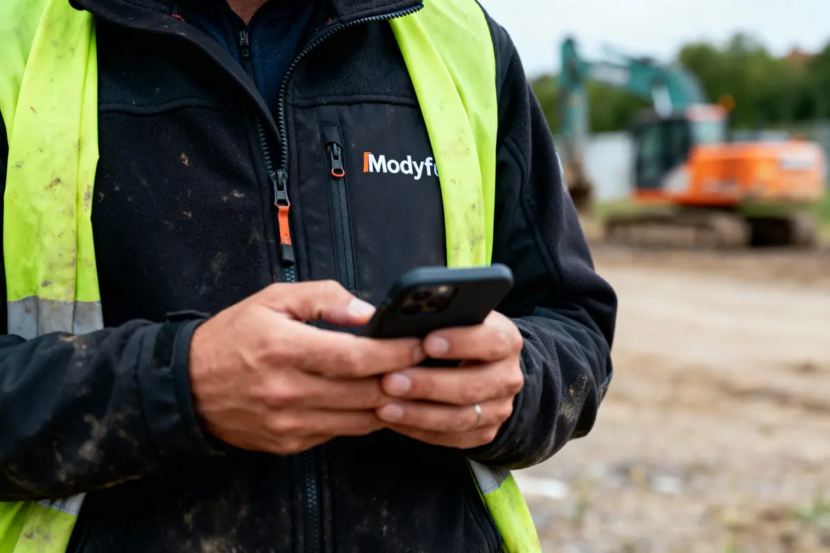 Homme en gilet fluorescent utilisant un smartphone sur un chantier avec une pelleteuse en arrière-plan.