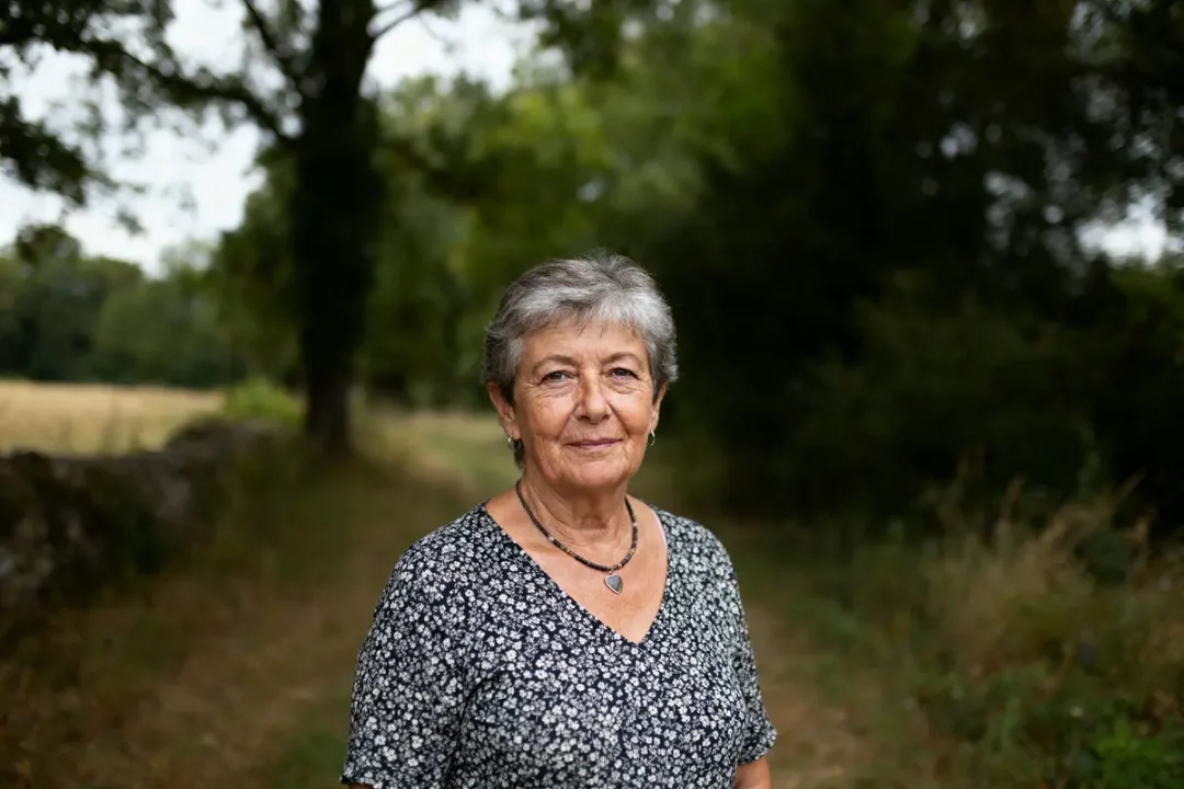 Une femme âgée debout dans un chemin forestier, portant une robe à motifs floraux.