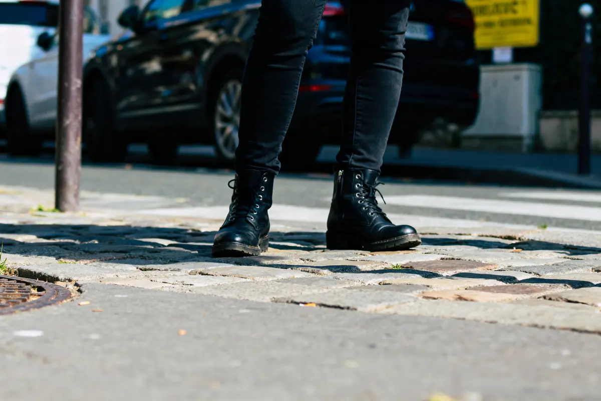 Personne portant des bottes noires sur un trottoir en ville, voitures garées en arrière-plan.