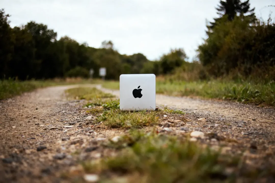 Mac Mini posé sur un chemin de gravier entouré de végétation, ciel légèrement nuageux en arrière-plan.
