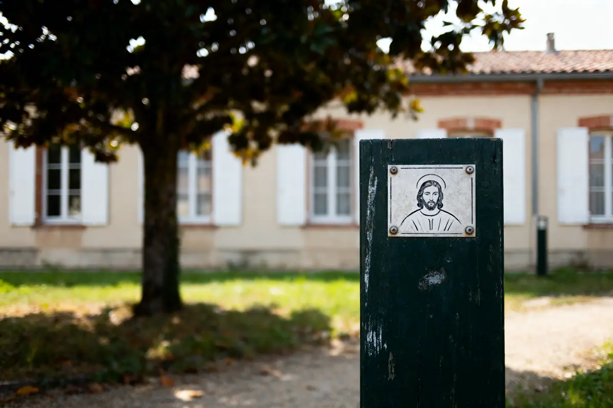 Plaque avec illustration religieuse sur poteau, devant maison avec fenêtres et volets, arbre en arrière-plan.