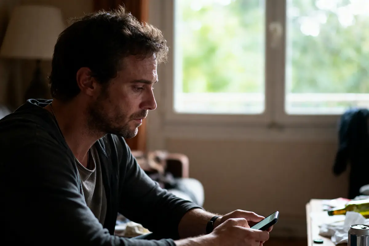 Homme concentré utilisant un smartphone, assis chez lui près d'une fenêtre. Éclairage intérieur tamisé, ambiance contemplative.