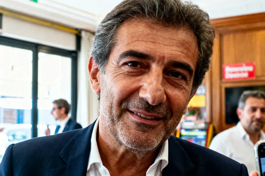 Homme souriant en costume sombre, intérieur lumineux, personnes floues en arrière-plan.