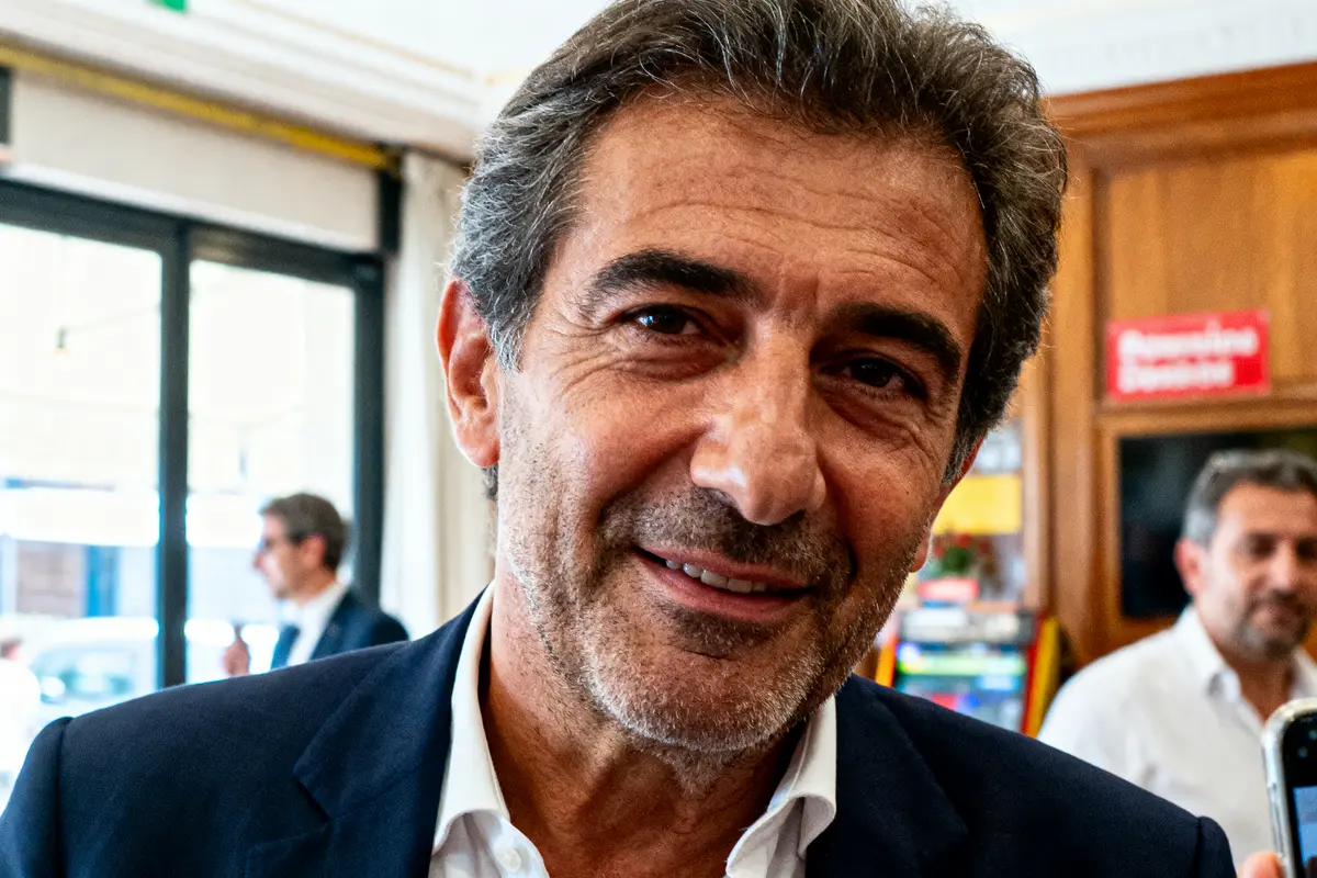 Homme souriant en costume sombre, intérieur lumineux, personnes floues en arrière-plan.