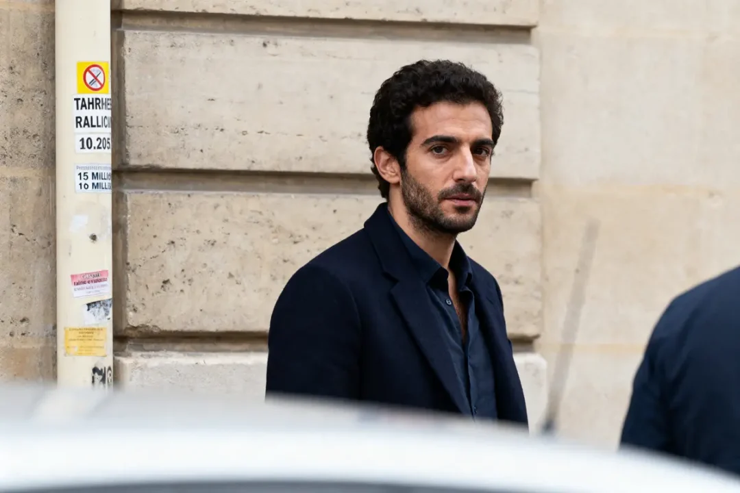 Homme en manteau foncé, rue parisienne, regarde vers la caméra, mur en pierre en arrière-plan.