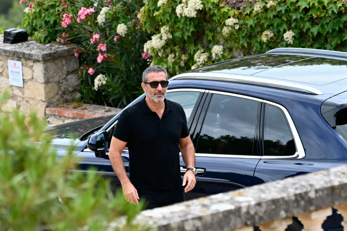Homme en noir avec lunettes de soleil, devant une voiture, fleurs et mur en arrière-plan.