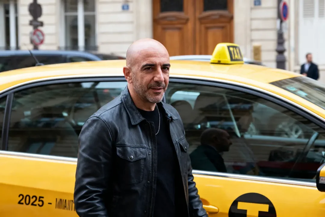Homme en veste noire devant un taxi jaune, bâtiments en arrière-plan.