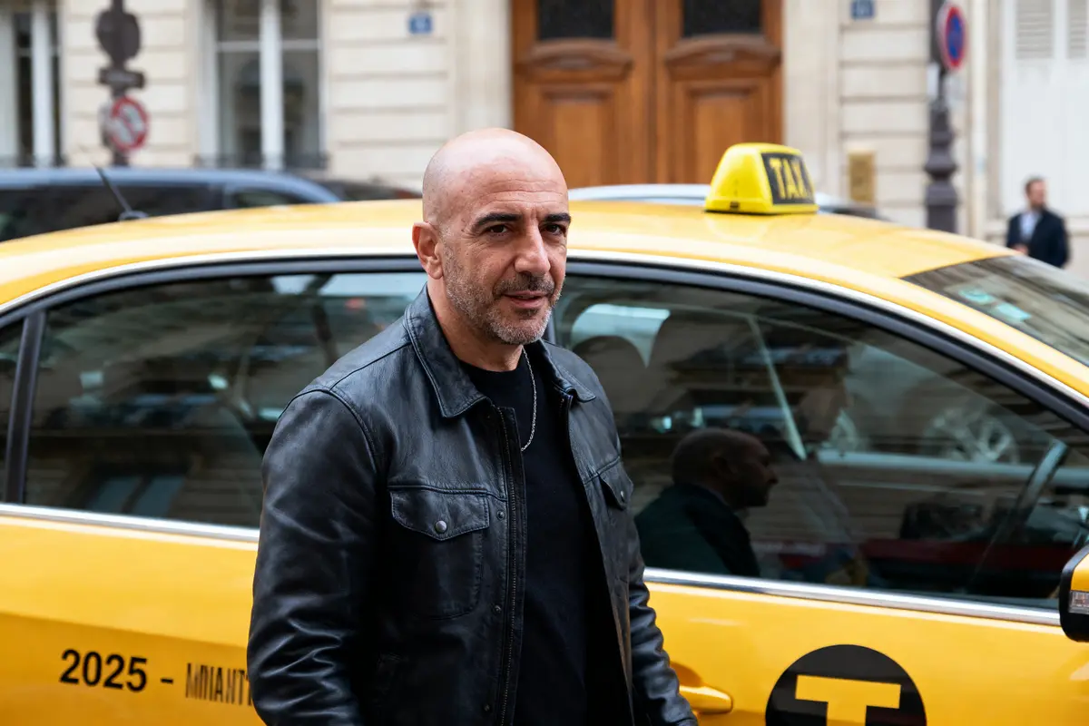 Homme en veste noire devant un taxi jaune, bâtiments en arrière-plan.