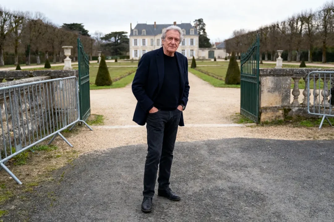 Homme debout devant des jardins soignés et un grand bâtiment en arrière-plan, portant une tenue sombre.