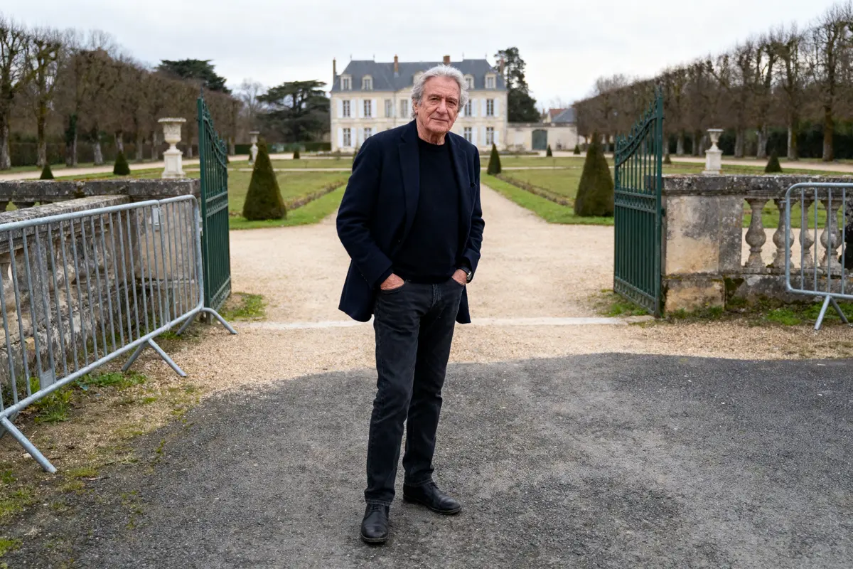 Homme debout devant des jardins soignés et un grand bâtiment en arrière-plan, portant une tenue sombre.