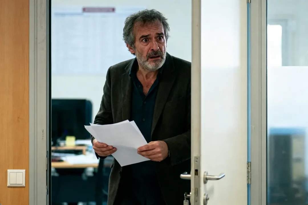 Homme barbu, expressions préoccupées, tenant des papiers, franchissant une porte de bureau.